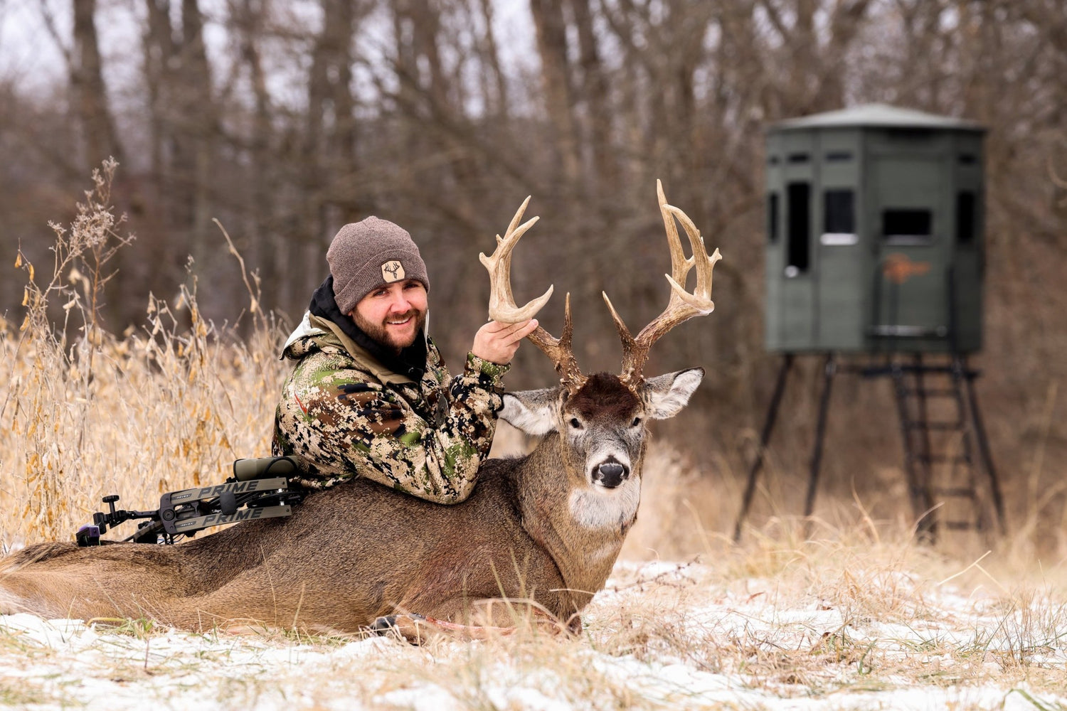 Hunter with a harvested whitetail buck in a snowy field, a 360 Pro elevated hunting blind on a tower stand in the background
