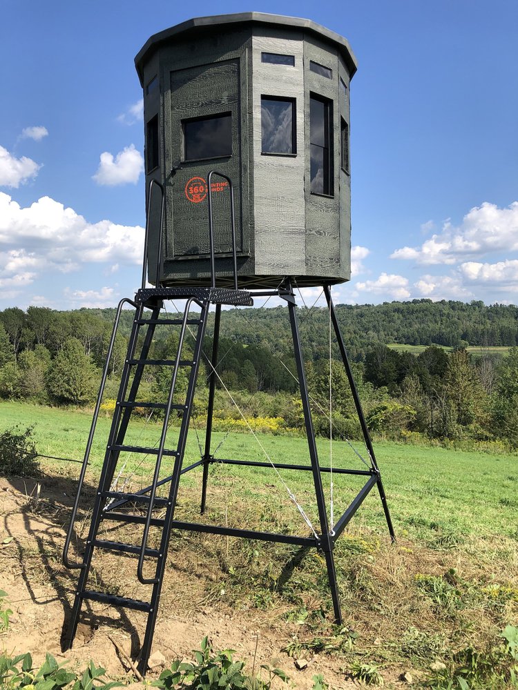360 Pro hunting blind on a tower stand with a ladder, set in a field under a blue sky with clouds